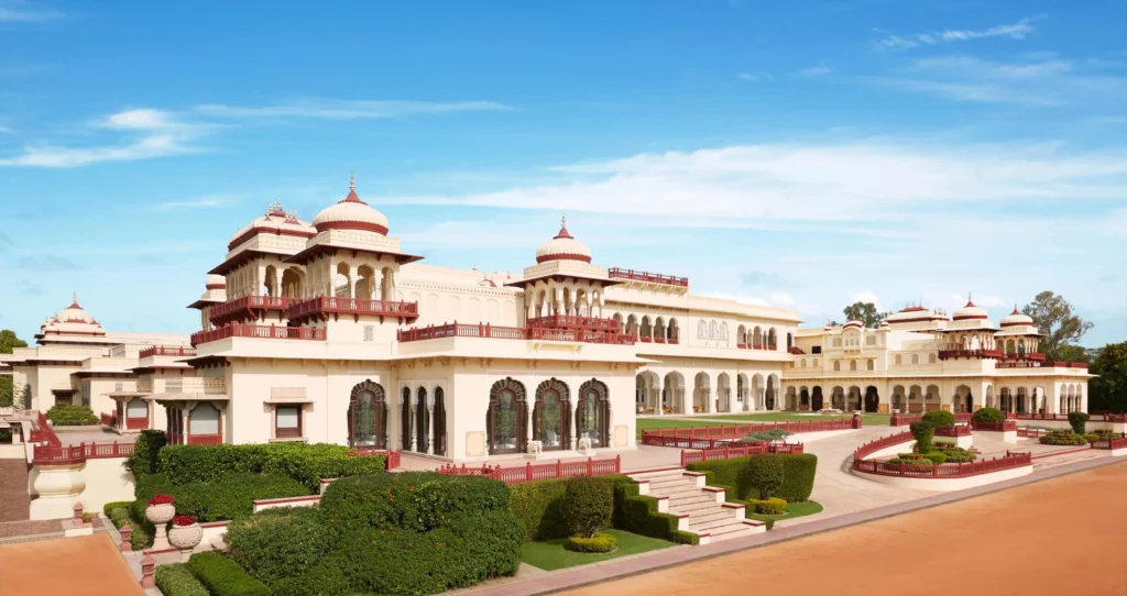 A wide-angle daytime shot of the Rambagh Palace exterior featuring its iconic yellow sandstone walls, red-domed chhatris, and sprawling green lawns under a bright blue sky.