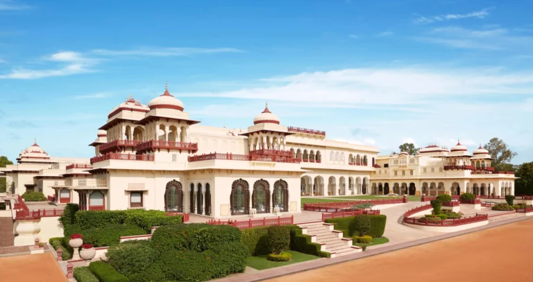A wide-angle daytime shot of the Rambagh Palace exterior featuring its iconic yellow sandstone walls, red-domed chhatris, and sprawling green lawns under a bright blue sky.