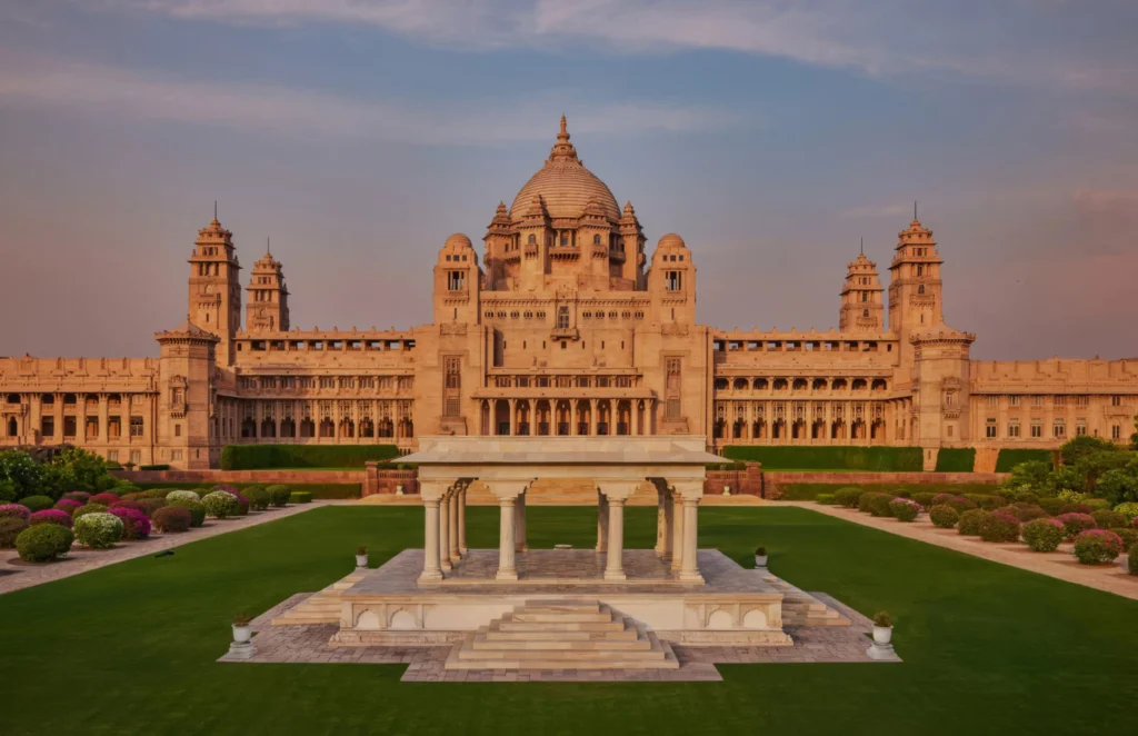 Exterior view of Umaid Bhawan Palace in Jodhpur featuring golden sandstone architecture, a central dome, and lush green gardens.