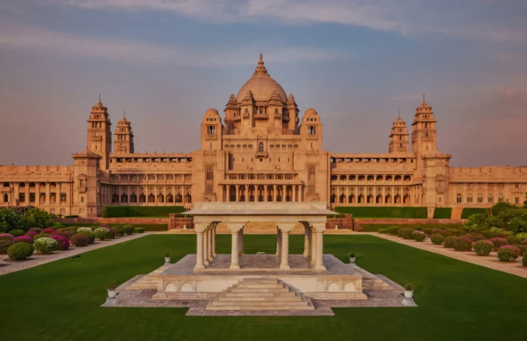 Exterior view of Umaid Bhawan Palace in Jodhpur featuring golden sandstone architecture, a central dome, and lush green gardens.